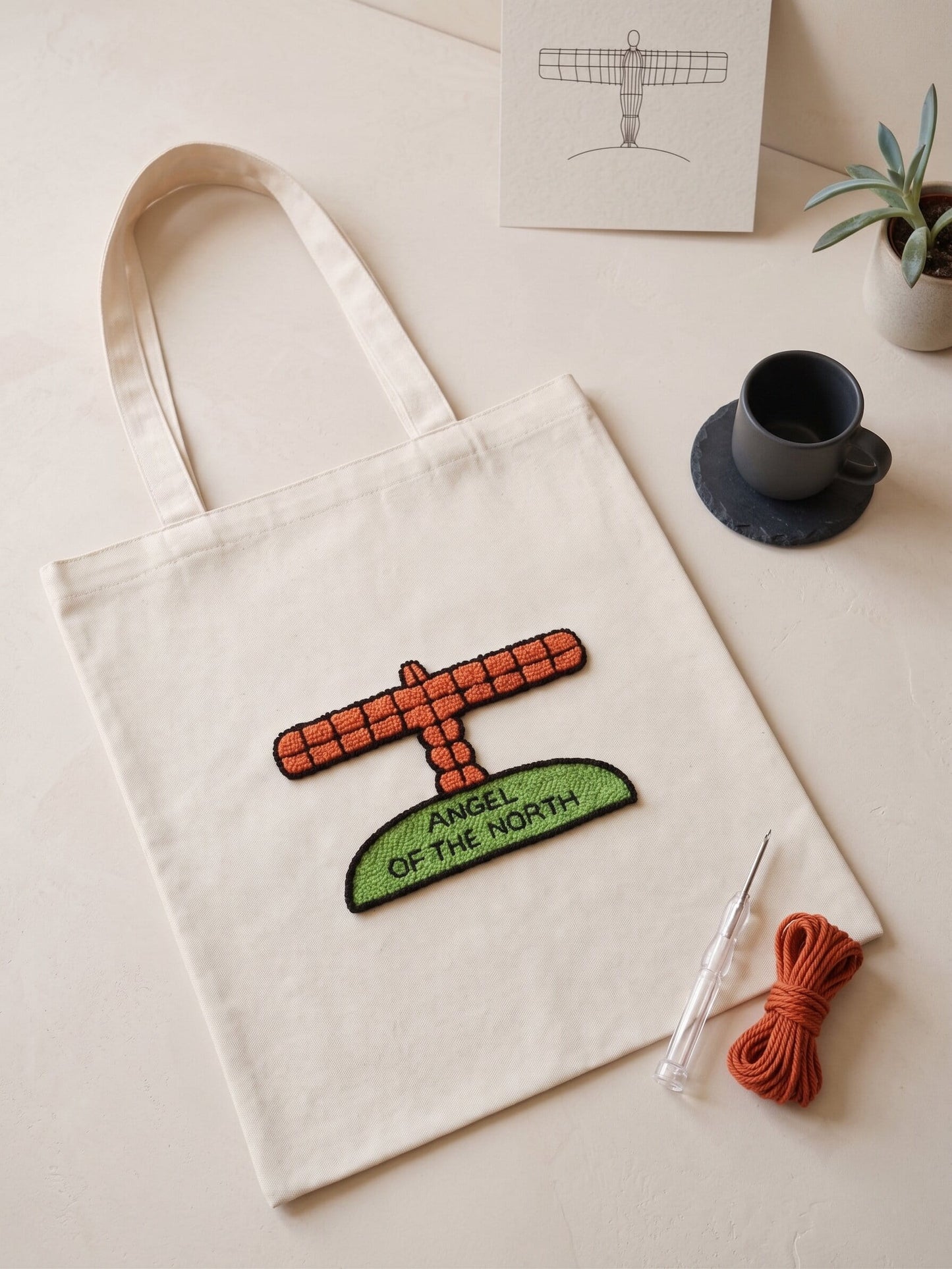 Angel of the North tote bag with handmade punch needle sculpture design and lettering, laid on a wooden table with yarn
