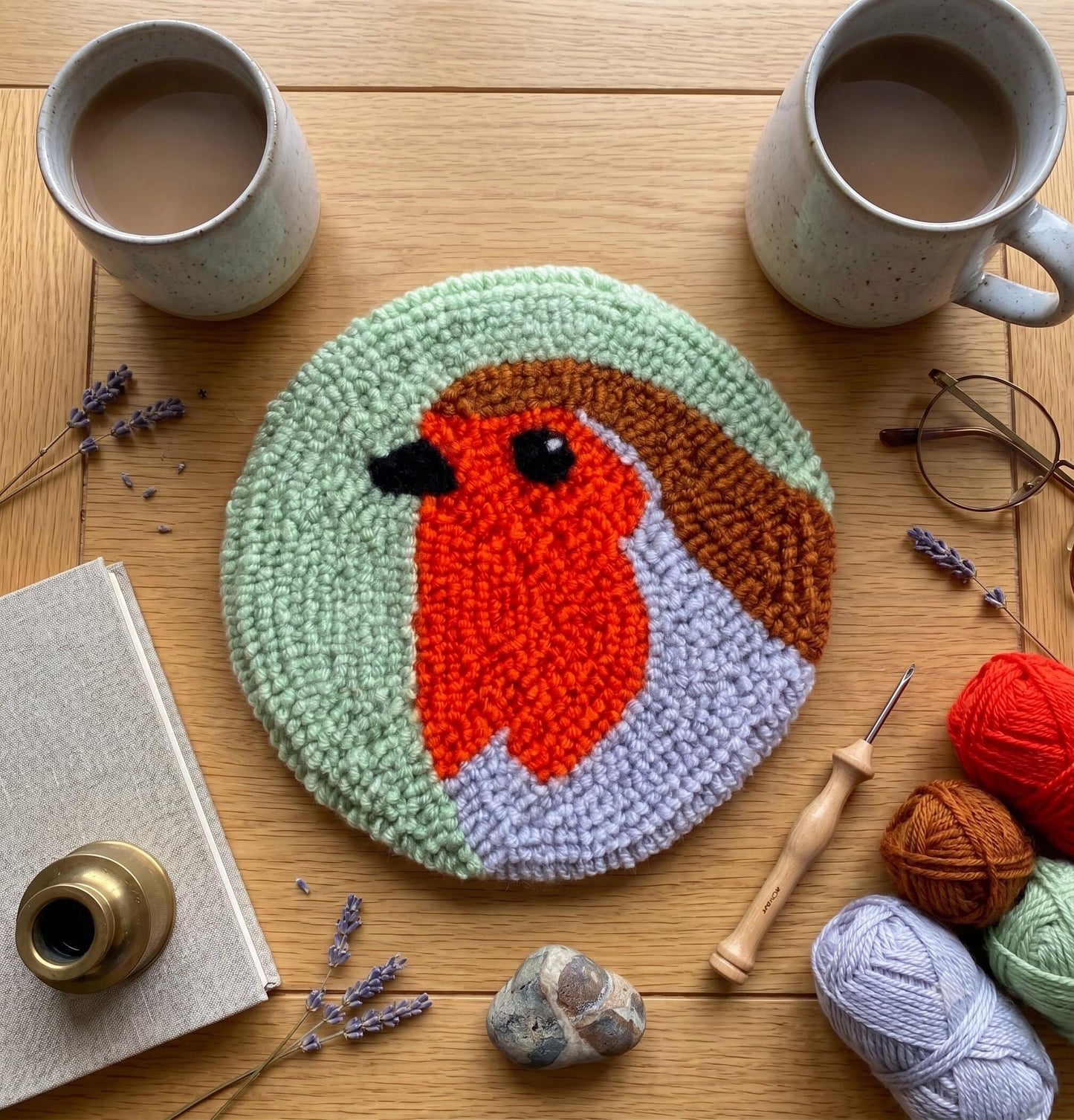 Round robin bird coaster with handmade punch needle design on sage green background, surrounded by mugs and yarn