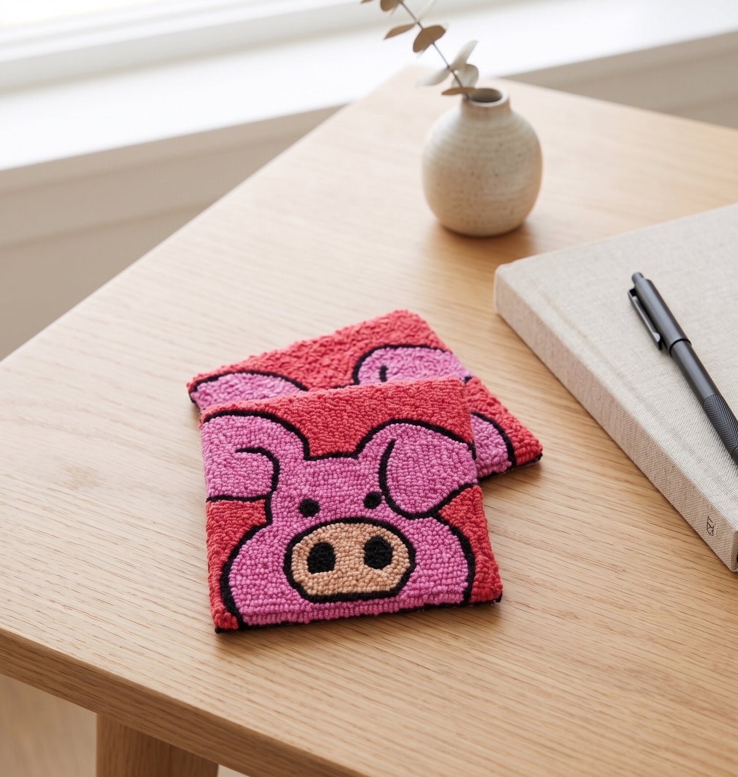 Pink pig coaster with handmade punch needle design, resting on a wooden desk next to a notebook and vase