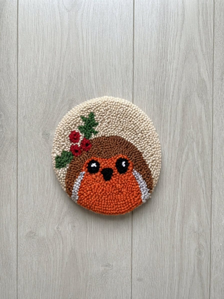A circular punch needle art piece featuring a cute robin bird with a green holly wreath, displayed on a light gray wood plank background.