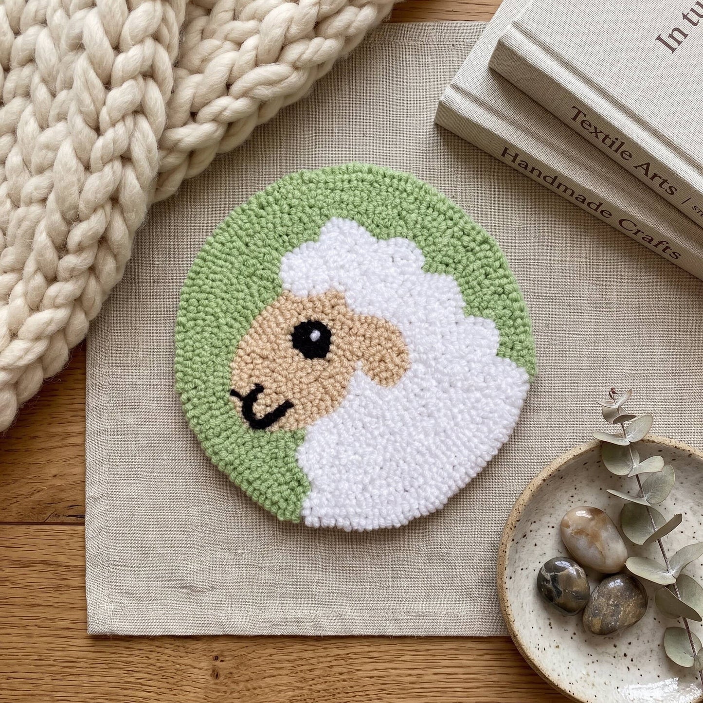 Round sheep coaster with handmade punch needle design on sage green background, surrounded by yarn and a wooden vase