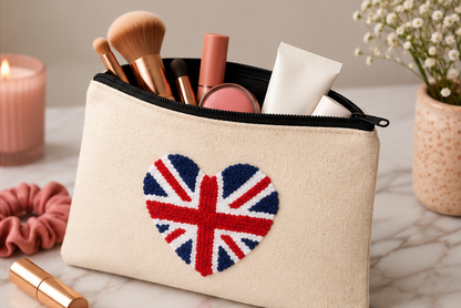 A beige canvas cosmetic pouch with a black zipper, featuring a red, white, and blue heart-shaped Union Jack design on the front. The pouch is shown open, filled with makeup items like brushes, a tube, and a compact, set on a marble surface with decorative props in the background.