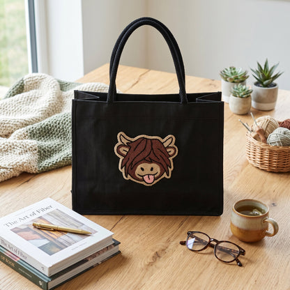 A black canvas tote bag with a cartoon cow character patch on the front, styled on a wooden table with a mug, glasses, books, and small potted succulents.