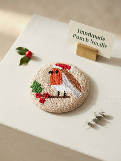 A round handcrafted punch needle pin featuring a robin bird with a Santa hat, displayed on a white surface next to a small sign that reads 'Handmade Punch Needle'.