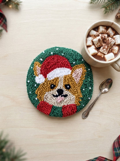A round punch needle holiday coaster featuring a corgi dog in a Santa hat, styled on a light wood tabletop next to a mug of hot chocolate with marshmallows and a spoon.