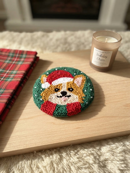 A round hand-tufted needlepoint coaster featuring a corgi dog in a Santa hat, styled on a wooden tray next to a plaid cloth and a candle.
