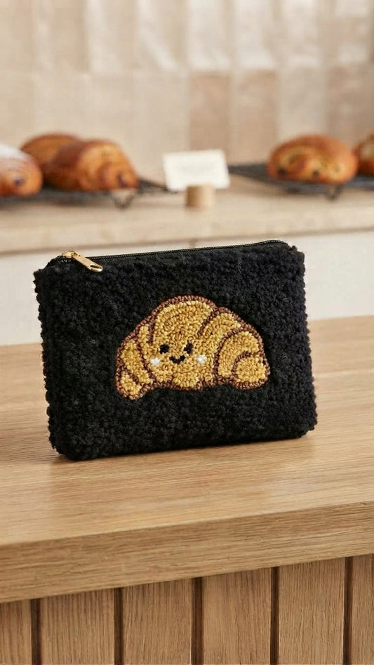 A black bouclé zip pouch featuring a smiling croissant applique on the front, resting on a wooden counter with a blurred bakery background.
