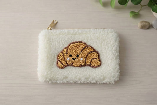 A white sherpa fleece zip pouch featuring a croissant-shaped beaded patch with a cute face, resting on a light wood surface with a gold zipper pull.