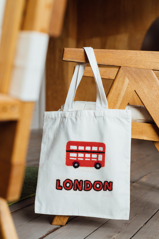 White tote bag with red London bus design hanging on a wooden chair.