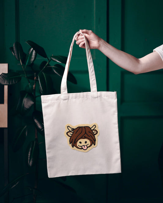 White tote bag with a highland cow design held by a person against a green background