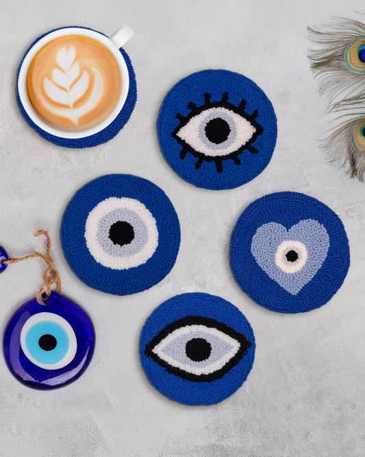 A set of round, blue coasters with white and black evil eye designs, displayed alongside a cup of coffee and a decorative blue Evil Eye item.