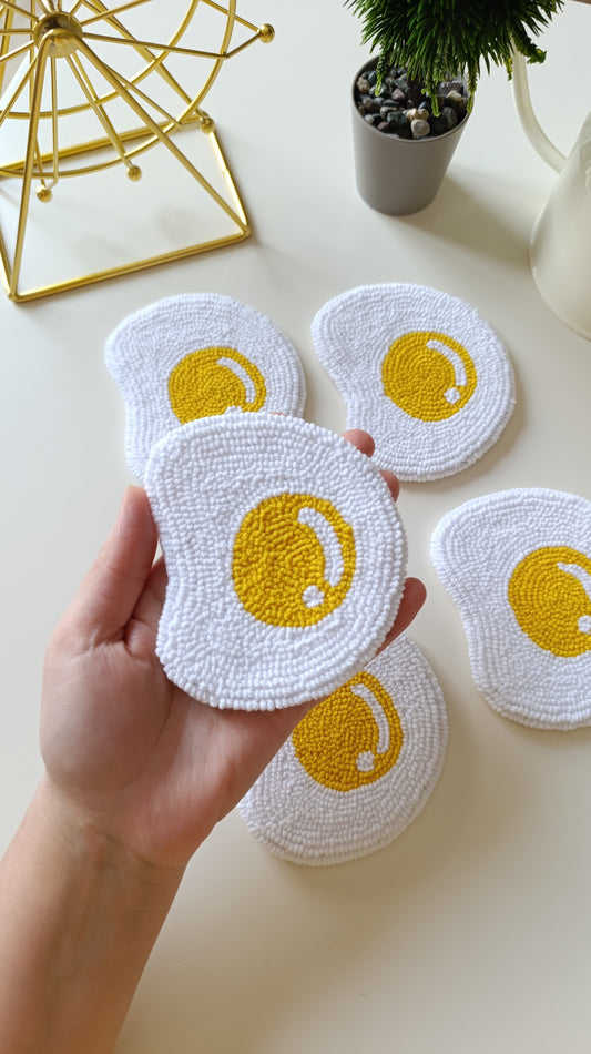 A hand holds a white textured dish towel with a yellow fried egg design, with four matching towels arranged on a light beige tabletop in the background.
