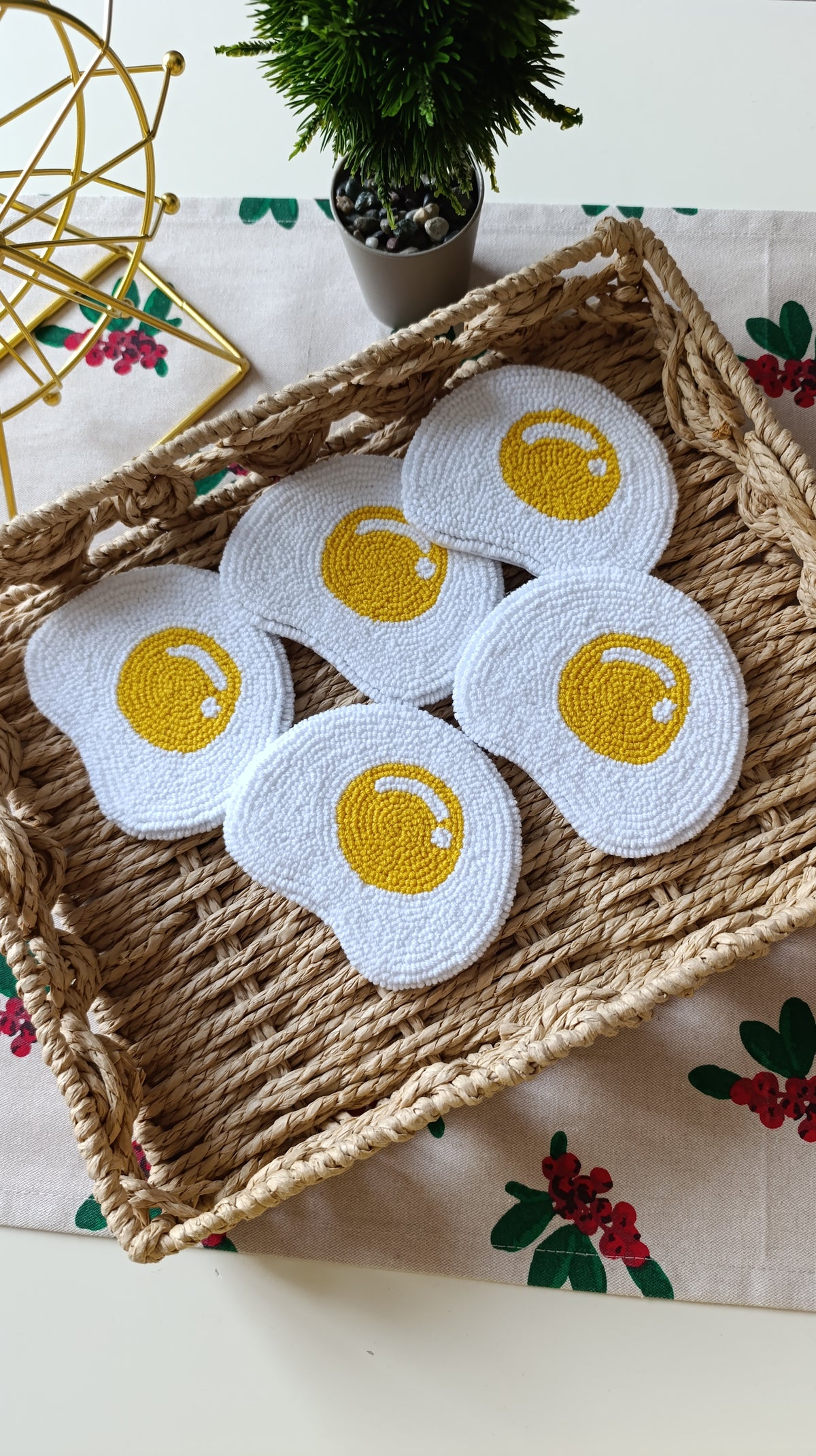 Five white crocheted coasters shaped like fried eggs with yellow centers, arranged in a woven straw tray on a floral tablecloth.