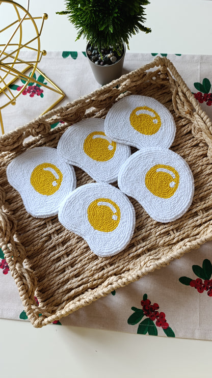 Five white crocheted coasters shaped like fried eggs with yellow centers, arranged in a woven straw tray on a floral tablecloth.