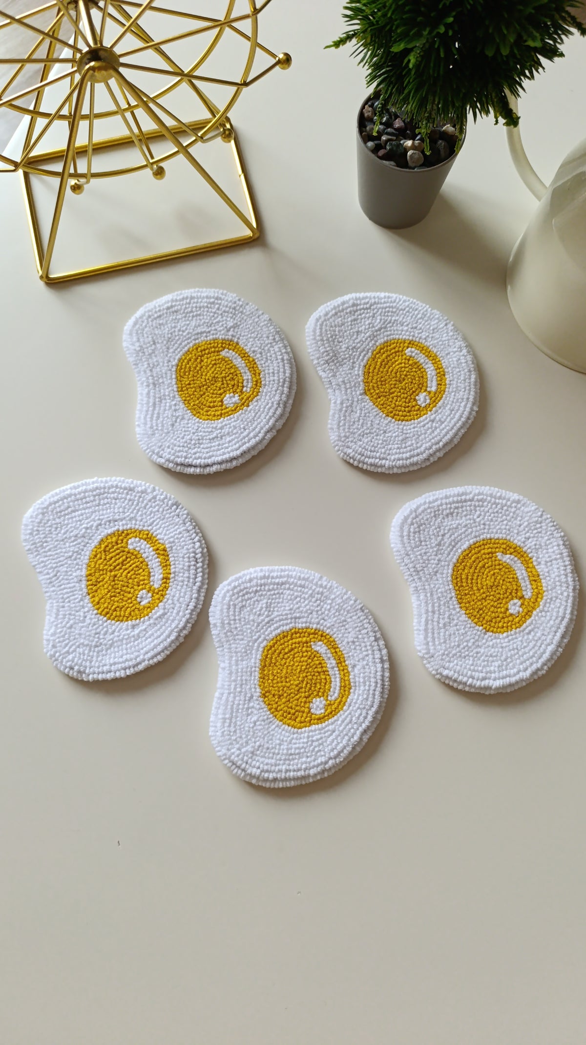 Five white textured dishcloths shaped like fried eggs with yellow yolks, arranged on a light beige tabletop with decorative props including a gold wire stand, a small potted plant, and a white pitcher.