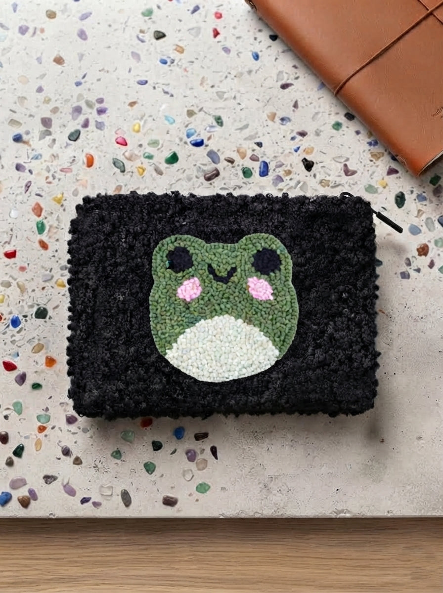 A black textured pouch with a green, white, and pink frog face design, displayed on a speckled beige surface with a brown leather strap in the corner.