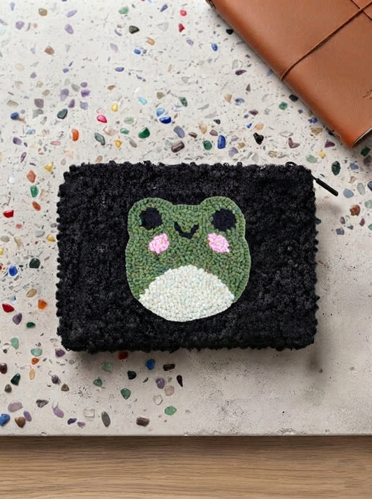 A black textured pouch with a green, white, and pink frog face design, displayed on a speckled beige surface with a brown leather strap in the corner.