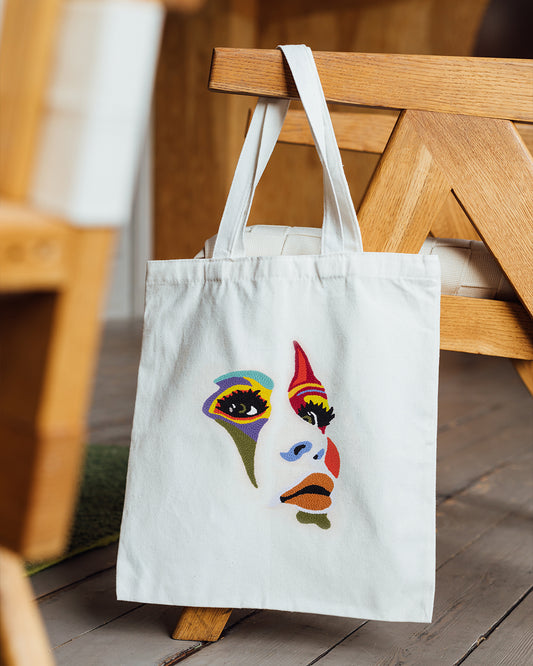 White tote bag with a colourful woman's face design hanging on a wooden chair