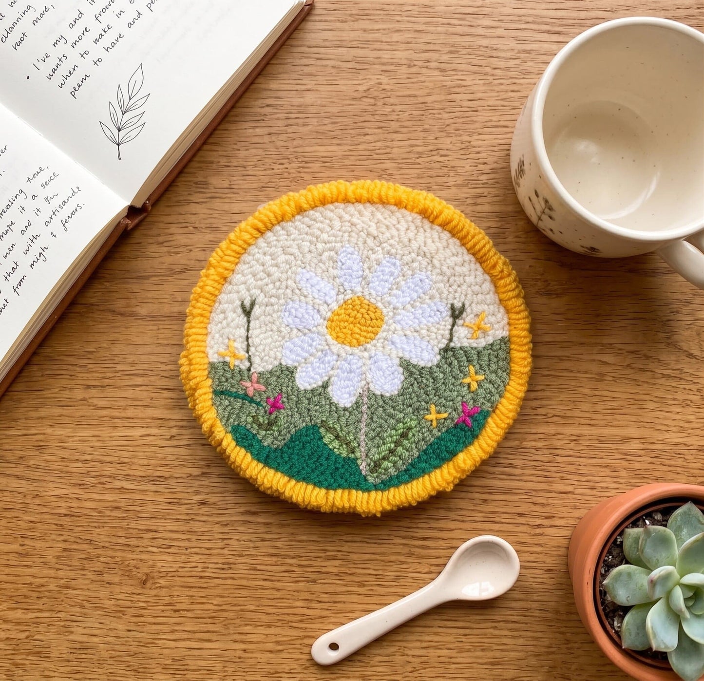 Handmade floral punch needle coaster with daisy design styled on a wooden table beside a cup, notebook, spoon, and succulent plant.