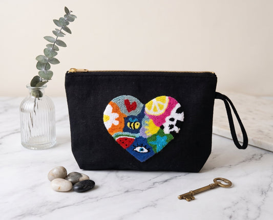Handmade punch needle black canvas clutch bag featuring a colourful patchwork heart design with mixed motifs including a bee, lemon slice, evil eye and watermelon, displayed on a marble surface with a vintage key and eucalyptus stems.