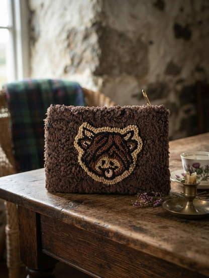 A brown textured clutch bag with a highland cow face motif in dark brown and beige, displayed on a rustic wooden table in a cozy, dimly lit room.