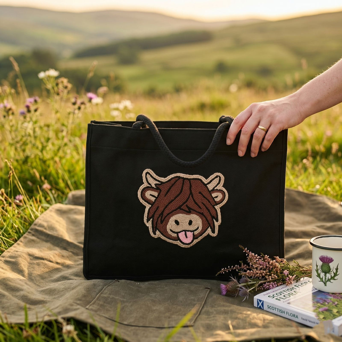 A black canvas tote bag with a cartoon Highland cow patch on the front, held by a person's hand in an outdoor grassy field at sunset. A book, flowers, and a mug sit on a blanket beside the bag.