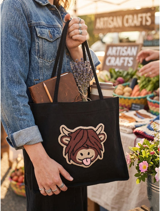 A person in a denim jacket holds a black canvas tote bag at an outdoor market. The bag features a cartoonish embroidered cow face with brown hair and a pink tongue.