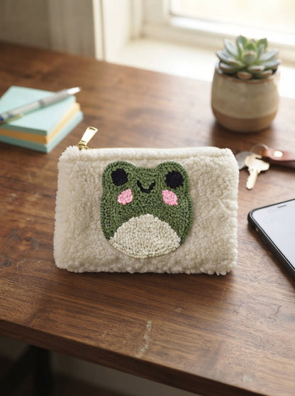 A cream-colored fuzzy pouch with a crocheted green frog face on the front, resting on a wooden desk next to a notebook, keys, and a smartphone.