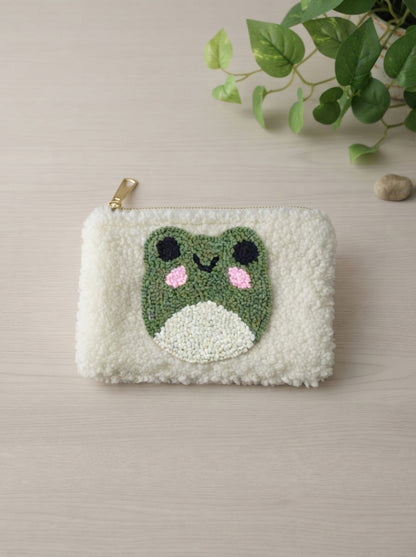 A cream-colored fluffy faux fur pouch with a green beaded frog face applique on the front, featuring a gold zipper pull, photographed on a light wood surface with a green plant in the background.