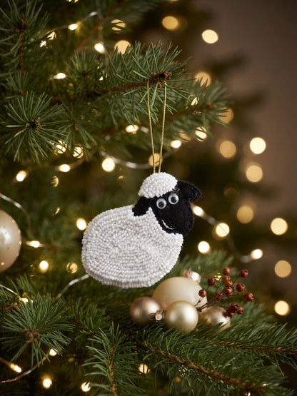 A sheep-shaped Christmas ornament with a white textured body and black face hangs on a decorated Christmas tree with warm string lights and gold ornaments.