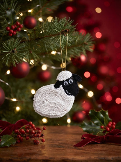 A handmade sheep-shaped Christmas ornament with a white textured body and black face hangs from a gold string on a decorated Christmas tree, with red ornaments, lights, and holly in the background.