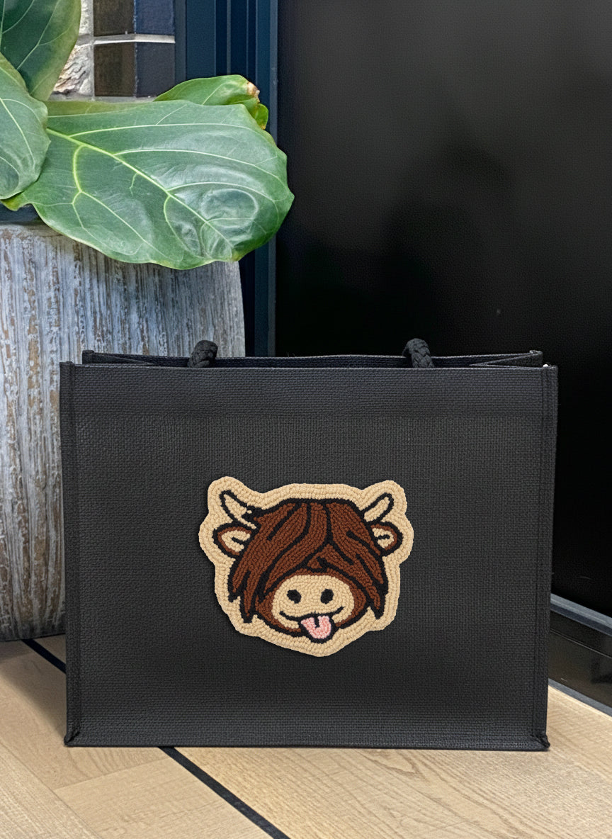 A black canvas tote bag with a cartoon cow patch on the front, placed on a light wood floor next to a potted green plant.