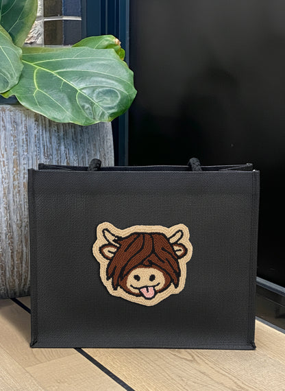 A black canvas tote bag with a cartoon cow patch on the front, placed on a light wood floor next to a potted green plant.