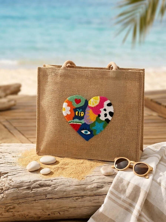 Handmade jute shoulder bag with colorful punch needle heart design placed on a beach setting with sand, sunglasses, and ocean in the background.