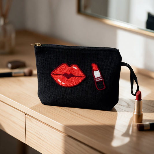 A black canvas cosmetic pouch with embroidered red lips and lipstick motifs, sitting on a light wood vanity table next to lipstick tubes.
