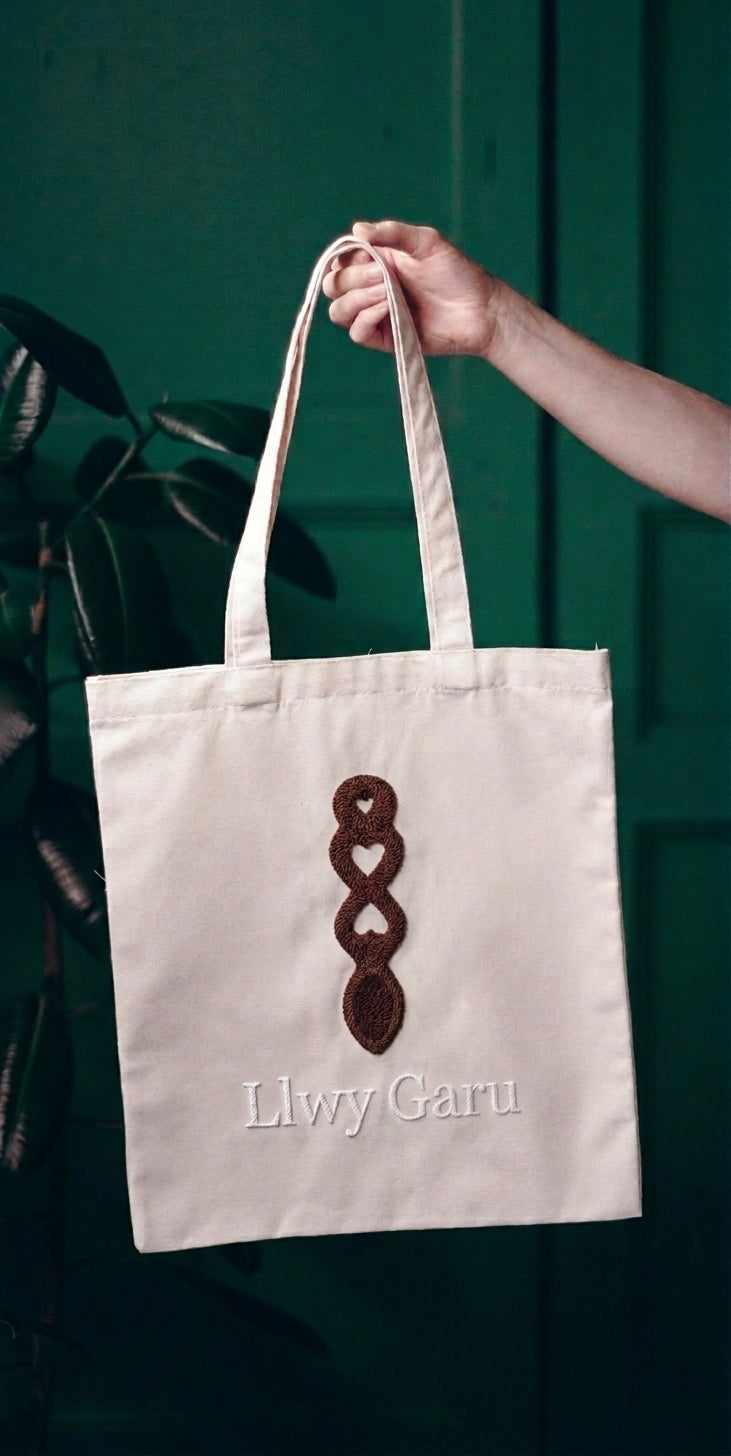 Welsh love spoon tote bag with handmade punch needle spoon design and Llwy Garu lettering, held by hand against a green door