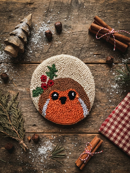 A round punch needle art piece of a robin bird with a holly wreath, displayed on a rustic wooden table surrounded by Christmas-themed props like pine branches, cinnamon sticks, and a red gingham cloth.