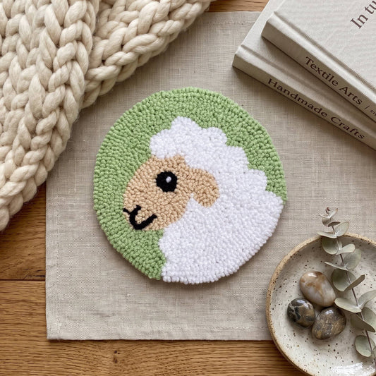 Round sheep coaster with handmade punch needle design on sage green background, surrounded by yarn and a wooden vase