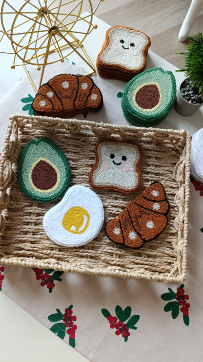 A set of cute, hand-knit breakfast-themed felt patches, including avocado, toast, croissant, and egg shapes, displayed on a woven tray over a floral tablecloth.