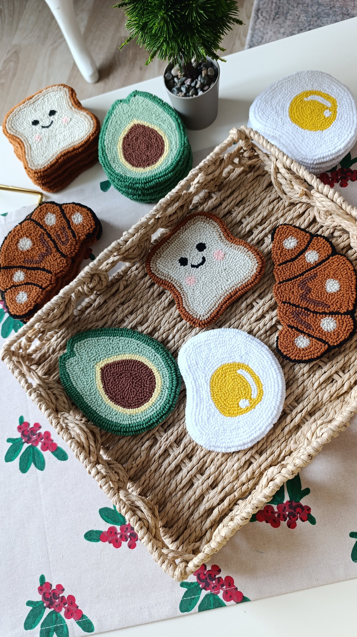 A styled flat-lay of crocheted or knitted breakfast-themed items, including avocado halves, toast slices with faces, croissants, and fried eggs, arranged on a woven tray and a floral tablecloth.