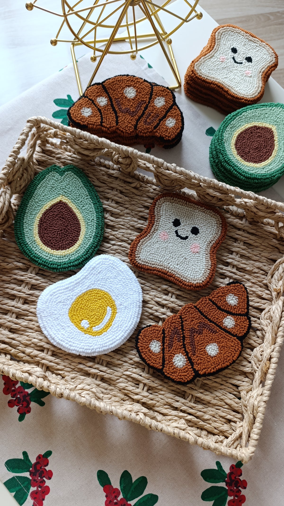 A set of crocheted or embroidered food-themed coasters or pads, including avocado, croissant, toast, and egg shapes, arranged in a woven tray on a floral tablecloth.