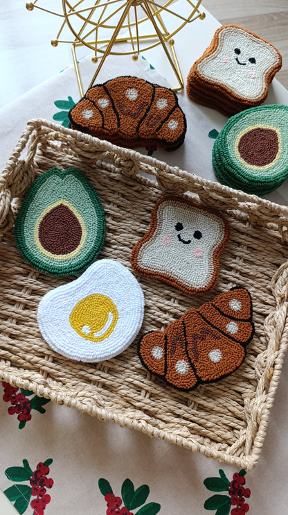 A set of crocheted or embroidered food-themed coasters or pads, including avocado, croissant, toast, and egg shapes, arranged in a woven tray on a floral tablecloth.
