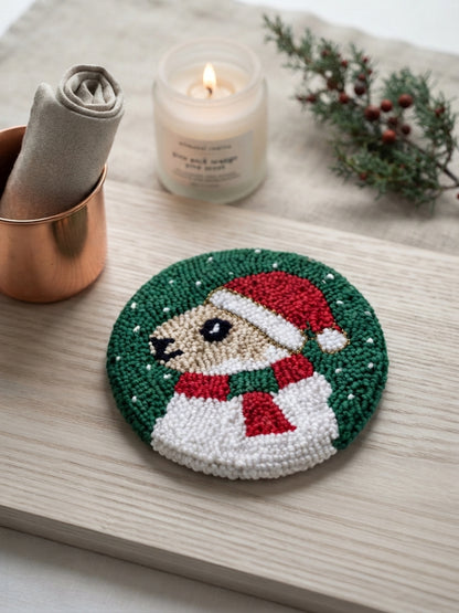 A round hand-punched bead art coaster featuring a dog in a Santa hat and scarf on a green background, styled on a light wood tray with a candle, a rolled napkin in a copper cup, and evergreen branches.