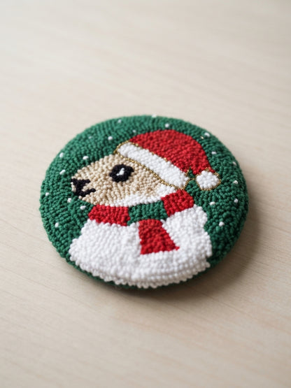A round hand-tufted decorative piece featuring a smiling dog wearing a red Santa hat and red-and-green scarf, set against a dark green textured background with small white dot details, photographed on a light wood surface.