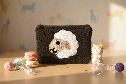 A dark brown textured pouch with a raised sheep face motif on the front, styled on a tabletop with macarons, a small cat figurine, and yarn balls against a softly patterned background.