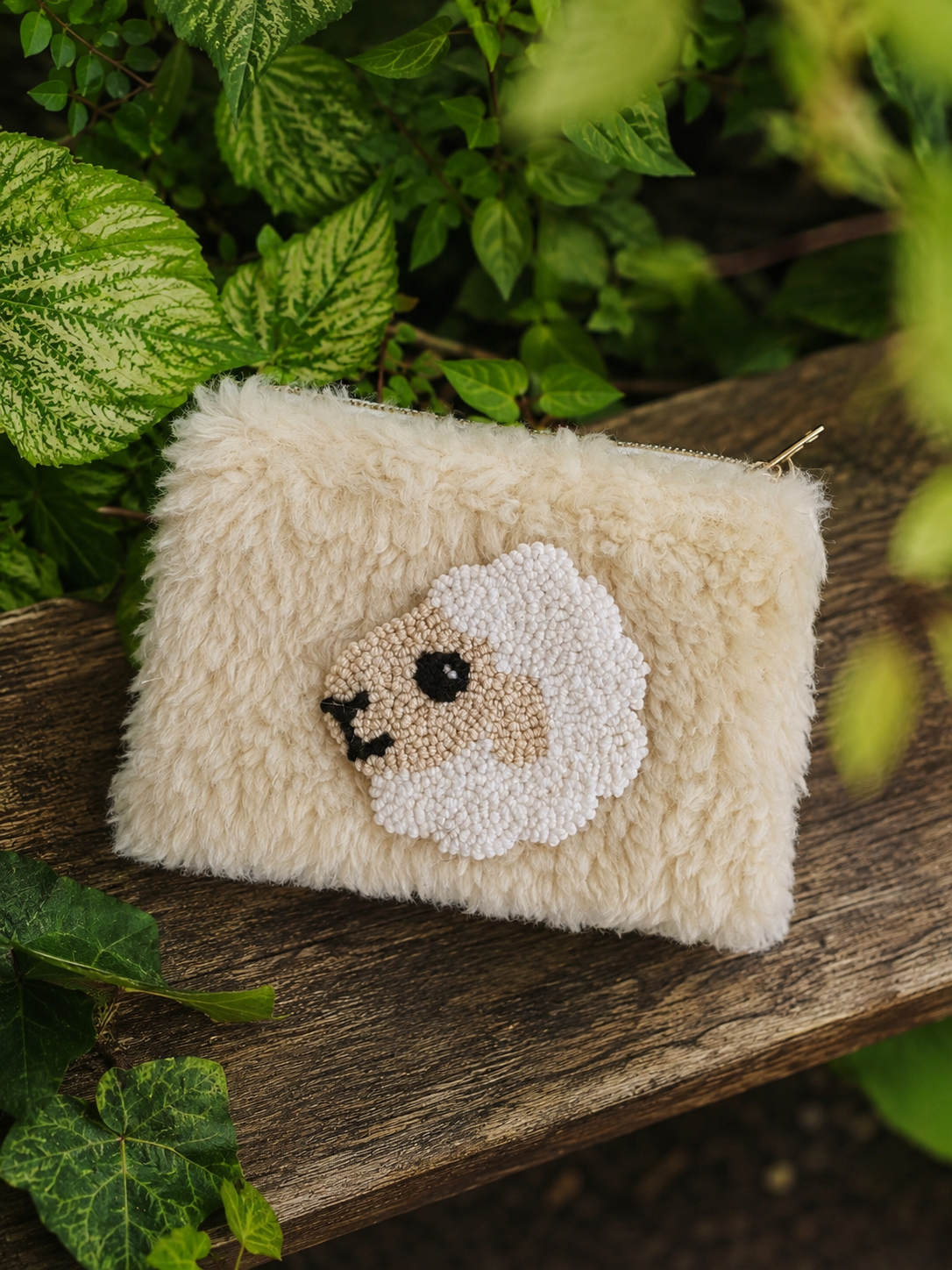 A small fluffy cream-colored pouch with a sheep face design, resting on a rustic wooden surface surrounded by green leaves.