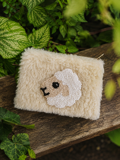 A small fluffy cream-colored pouch with a sheep face design, resting on a rustic wooden surface surrounded by green leaves.