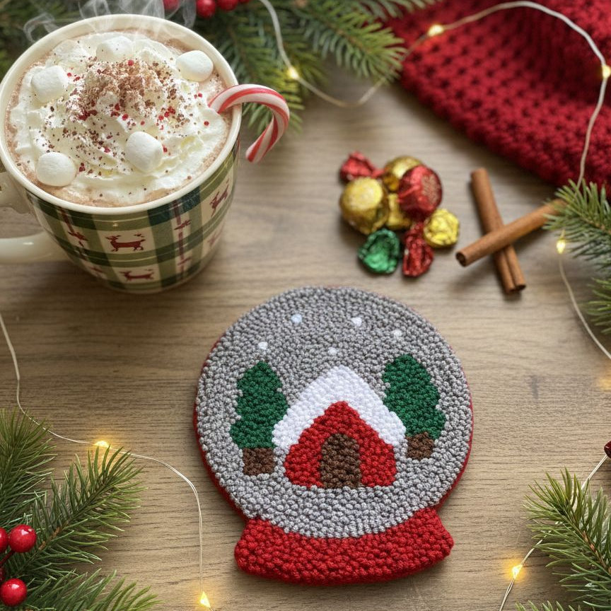Handcrafted snow globe punch needle coaster with a mug of hot chocolate on a wooden table.