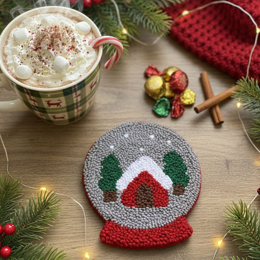 Handcrafted snow globe punch needle coaster with a mug of hot chocolate on a wooden table.