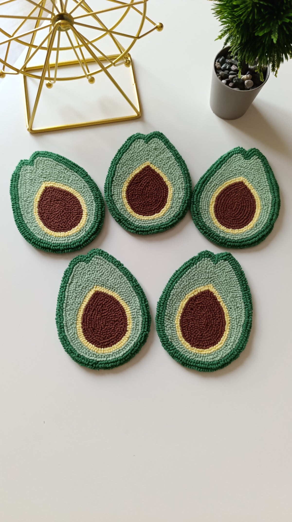 Five avocado half-shaped coasters arranged on a white surface, with a gold geometric decor object and a small potted plant in the background.